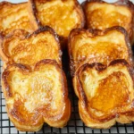 Golden croissant bread French toast slices arranged neatly on a black wire rack over a tray.