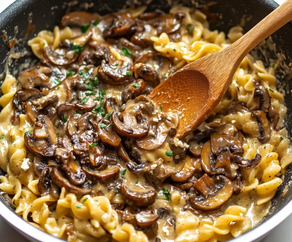 A skillet filled with freshly cooked caramelized mushroom and goat cheese pasta, stirred with a wooden spoon, with creamy sauce coating the pasta.