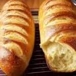 Freshly baked homemade French bread loaves cooling on a wire rack, with a crispy golden crust and soft interior, one loaf partially torn to reveal texture.